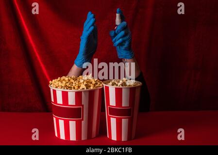 Ansicht der Frau in Latexhandschuhen, die Händedesinfektionsmittel in der Nähe von Eimern mit Popcorn auf roter Oberfläche mit Velours im Hintergrund hält Stockfoto