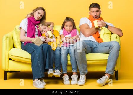 Kranke Familie mit Kindern in Schals hält Tassen mit heißen Getränken während auf dem Sofa auf gelb sitzen Stockfoto