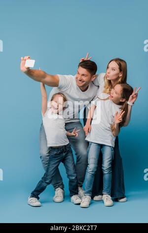 Fröhliche Familie zeigt Siegeszeichen während Selfie auf Smartphone auf blau Stockfoto