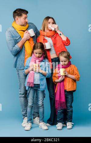 Traurig kranke Familie mit Kindern in Schals halten Tassen mit heißen Getränken auf blau Stockfoto