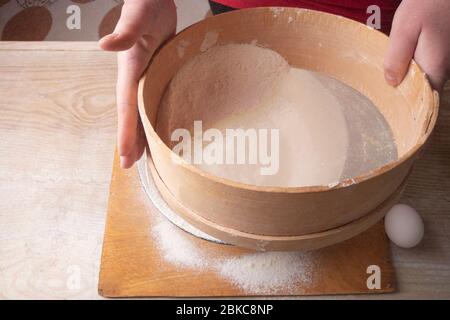 Große Schüssel, Schneidebrett, Sieb zum Sieben von Mehl auf dem Tisch. Weibliche Hände sieben Mehl in der Küche. Stockfoto