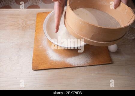 Große Schüssel, Schneidebrett, Sieb zum Sieben von Mehl auf dem Tisch. Weibliche Hände sieben Mehl in der Küche. Stockfoto