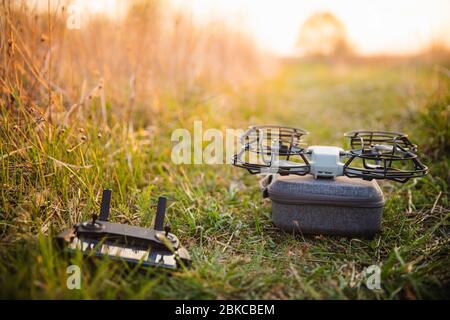 Kleine Drohne mit Propellerschutz auf grauem Gehäuse mit Steuertelefon auf grünem Frass im Sommerfeld mit warmem Sonnenuntergangslicht Stockfoto