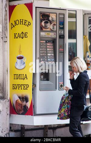 Lady nimmt sich unterwegs einen schnellen Kaffee aus einem Verkaufsautomaten im Freien auf den Straßen von Plovdiv, Bulgarien Stockfoto