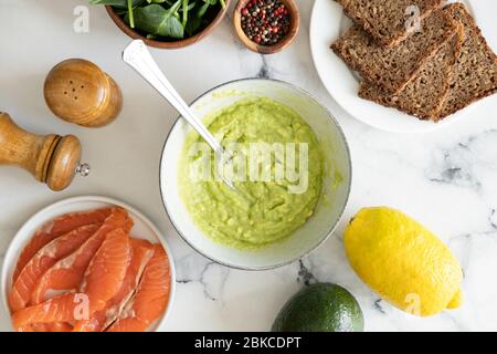 Avocado-Sauce in Schüssel und Zutaten für ein gesundes leckeres Sandwich mit Roggenbrot, geräuchertem Lachs Fisch, Avocado-Püree und Baby-Spinat-Blatt Stockfoto