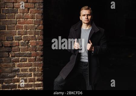 Junger ernsthafter Mann im Mantel, der neben der Wand steht und seinen Mantelkragen hält Stockfoto