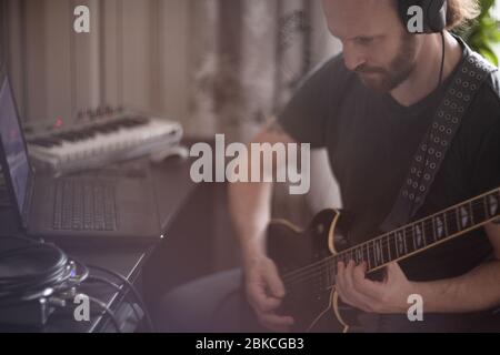Bärtiger Mann Musiker Musik spielen und komponieren ein Lied mit E-Gitarre Klavier und Laptop-Computer, während im Wohnzimmer sitzen, Corona Virus quar Stockfoto