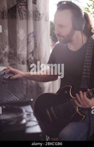 Bärtiger Mann Musiker Musik spielen und komponieren ein Lied mit E-Gitarre Klavier und Laptop-Computer, während im Wohnzimmer sitzen, Corona Virus quar Stockfoto