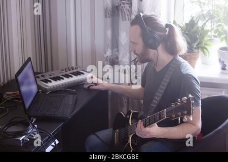 Bärtiger Mann Musiker Musik spielen und komponieren ein Lied mit E-Gitarre Klavier und Laptop-Computer, während im Wohnzimmer sitzen, Corona Virus quar Stockfoto