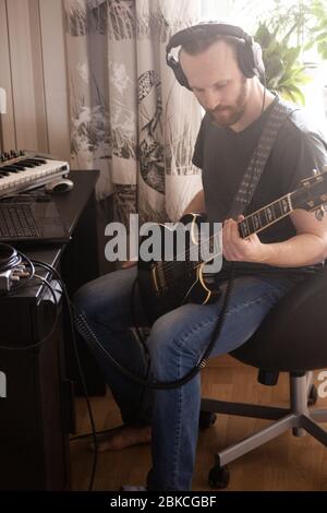 Bärtiger Mann Musiker Musik spielen und komponieren ein Lied mit E-Gitarre Klavier und Laptop-Computer, während im Wohnzimmer sitzen, Corona Virus quar Stockfoto