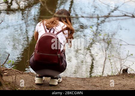 Rotschopf Mädchen mit einem Rucksack-Tasche hockt auf dem Flussufer. Erholung im Freien im Frühling Stockfoto