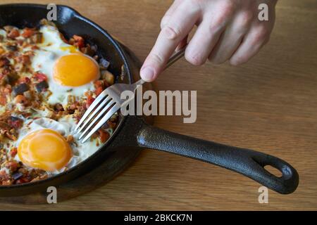 Hintergrund der Speisen, Spiegeleier mit Gemüse in der Pfanne und männliche Hand mit Gabel auf Holztisch, selektive Fokussierung Stockfoto
