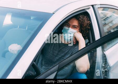 Ein junger Mann in einer medizinischen Maske, einem grauen T-Shirt und Jeans sitzt am Steuer eines Autos. Ein Mann in einer Schutzmaske fährt ein Auto. Schutz vor CO Stockfoto
