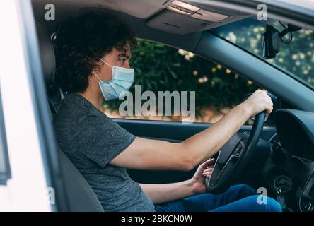 Junger Mann, der ein Auto in einer medizinischen Maske und grauem T-Shirt fährt. Mann in Schutzmaske sitzt am Steuer eines Autos. Schutz vor Coronavirus-Epid Stockfoto