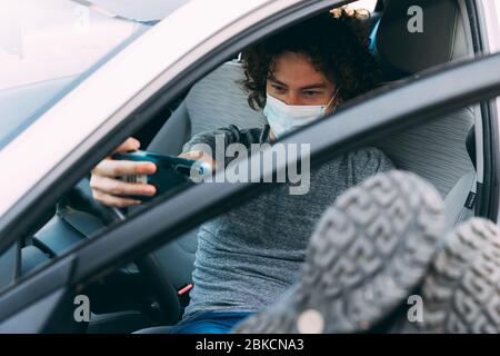 Der junge, lockige Mann sitzt im Auto mit seinen Beinen oben. Graues T-Shirt und blaue Jeans. Nimmt Selfie auf dem Mobiltelefon auf. Kerl in Schutzmaske. Coronavi Stockfoto