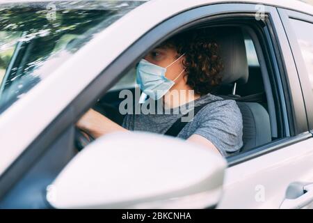 Ein Mann fährt ein Auto in einer medizinischen Maske. Ein Mann in einer Schutzmaske sitzt hinter dem Lenkrad eines Autos. Taxifahrer in Maske. Schutz vor Coronavirus-epid Stockfoto