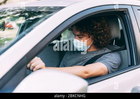 Ein Mann fährt ein Auto in einer medizinischen Maske. Ein Mann in einer Schutzmaske sitzt hinter dem Lenkrad eines Autos. Taxifahrer in Maske. Schutz vor Coronavirus-epid Stockfoto