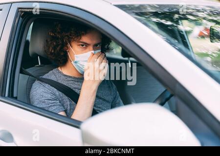 Ein Mann fährt ein Auto in einer medizinischen Maske. Ein Mann in einer Schutzmaske sitzt hinter dem Lenkrad eines Autos. Taxifahrer in Maske. Schutz vor Coronavirus-epid Stockfoto