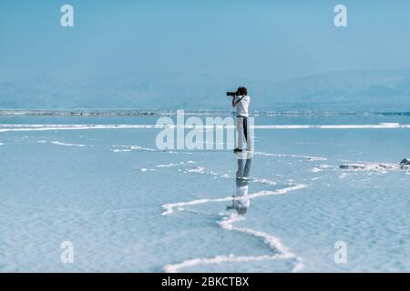 Der junge Mann mit lockigen Haaren fotografiert auf einer kleinen Insel im Toten Meer auf Teleobjektiv und spiegelt sich im Wasser. Wasser und Stockfoto