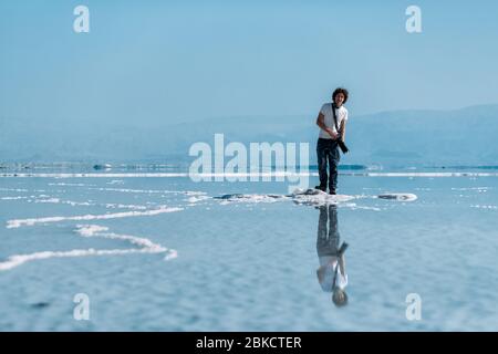 Ein junger Mann mit lockigen Haaren mit Kamera und Teleobjektiv, geht auf kleinen Inseln im Toten Meer und spiegelt sich im Wasser. Wasser und blauer Himmel Stockfoto