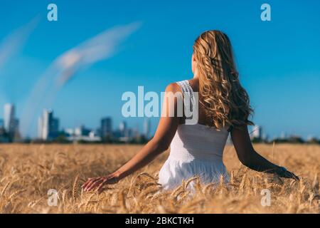 Junge blonde, lockige Frau von hinten in einem weißen Kleid in einem Weizenfeld vor blauem Himmel, verschwommene Stadtgebäude im Hintergrund. Frau berührt a s Stockfoto