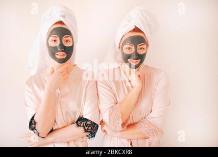 Zwei junge Schwestern in Bademänteln mit Handtüchern auf dem Kopf isoliert auf weißem Hintergrund. Spa zu Hause. Sie machen eine Maske für die Hautpflege auf der Stockfoto