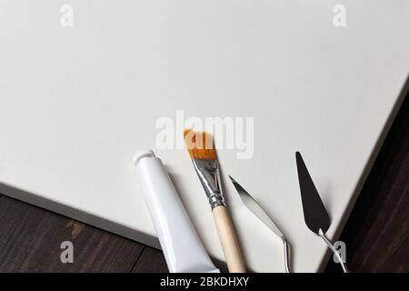 Ein weißer, blank getupfter Baumwollstoff auf Trage, Palettenmessern, synthetischem Pinsel und einem Rohr mit Öl- oder Acrylfarbe auf einem braunen Holztisch Stockfoto