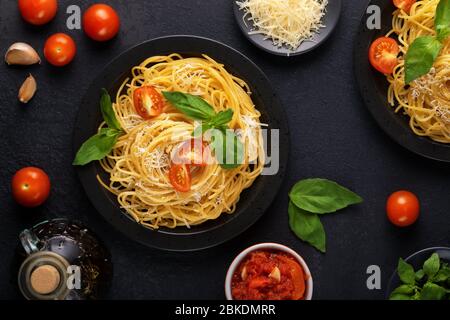 Zwei schwarze Teller mit vegetarischer, appetitlich-klassischer italienischer Spaghetti-Pasta mit Basilikum, Tomatensauce, Parmesan und Olivenöl auf einem dunklen Tisch. Draufsicht Stockfoto