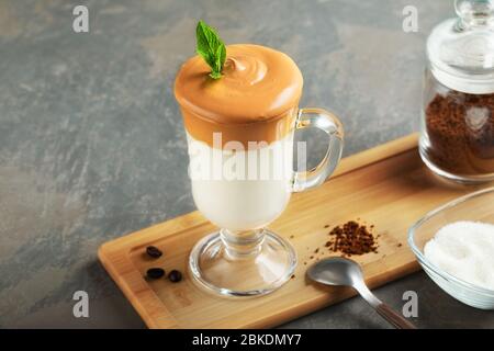 Dalgona schaumig Kaffee aus Glas auf Holzbrett auf grauem Hintergrund. Trend koreanisch Iced Latte Kaffee Getränk mit Schaum von Instant-Kaffee mit Zutaten. Stockfoto