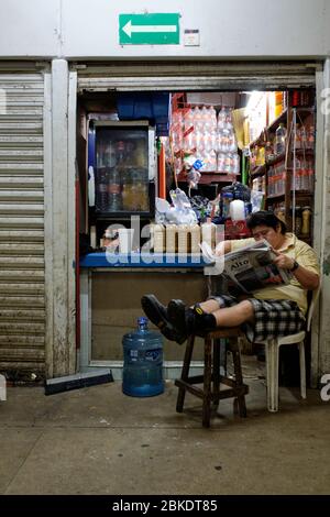 Mann, der die Zeitung liest, sitzt mit ausgestreckten Beinen auf einem Hocker vor einem kleinen Laden auf dem städtischen Markt von Merida. Stockfoto