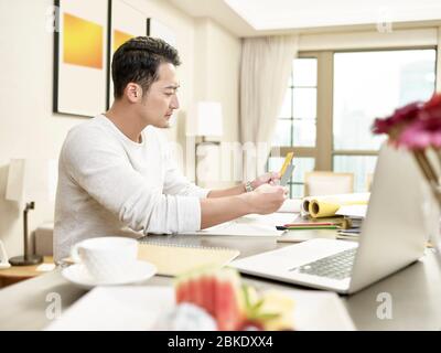Junge asiatische Mann Design professionelle Arbeit von zu Hause aus sitzen an der Küchentheke Vergleich zwei Farbkarten (Kunstwerk im Hintergrund digital altere Stockfoto