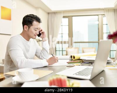 Junge asiatische Mann Design professionelle Arbeit von zu Hause aus sitzen in der Küche sprechen auf Handy (Kunstwerk im Hintergrund digital verändert) Stockfoto