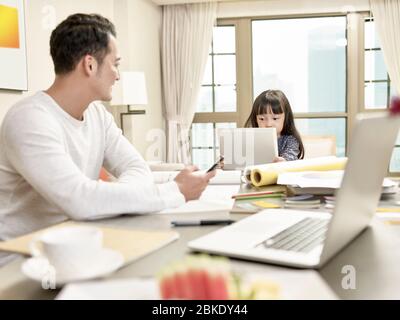 Junge asiatische Geschäftsmann kümmert sich um Tochter während der Arbeit von zu Hause (Kunstwerk im Hintergrund digital verändert) Stockfoto