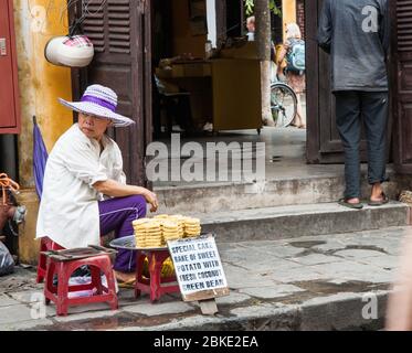 Hoi an Vietnam Oktober 17 2013; Frau Straße Verkäufer trägt modernen Hut sitzt im Hocker außerhalb Eingang zu einem Gebäude Verkauf Kuchen auf einem Tablett Stockfoto