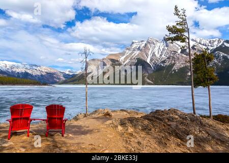 Rote Adirondack Stühle und schneebedeckte Berggipfel Landschaft. Lake Minnewanka, Frühfrühling Tag im Banff National Park, Rocky Mountains Alberta Kanada Stockfoto