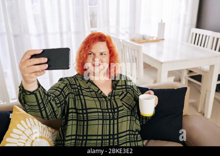 Lächelnde junge Frau im karierten Hemd, die mit einer Tasse Kaffee auf der Couch sitzt und Selfie nimmt Stockfoto