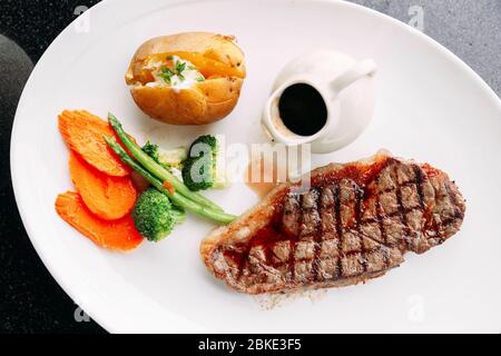 Draufsicht auf Holzkohle gegrilltes, knochenloses, trocken gereiftetes Top Loin Steak mit BBQ-Sauce, gebratenem Gemüse und gebackenen Kartoffeln auf einem weißen Teller. Stockfoto