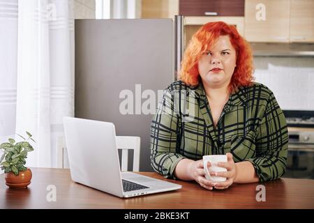 Porträt von hübschen plus size junge Frau sitzt am Küchentisch mit geöffneten Laptop, Kaffee trinken und Blick auf Kamera Stockfoto
