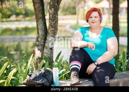 Ziemlich junge Frau plus Größe, die im Park ausruht und Musik in ihren Kopfhörern nach Joggin hört Stockfoto