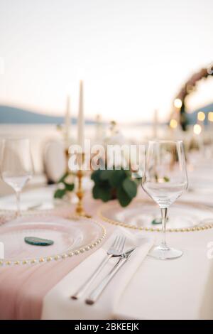 Nahaufnahme des Hochzeitstisch. Schöne Glaswaren - Teller mit goldenen Perlen, Weingläser, Messer und Gabeln, auf Hintergrund von Kerzen und Blumen Stockfoto