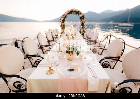 Nahaufnahme eines Festes am Esstisch für die Hochzeit. Ein Tisch steht am Strand mit Blick auf die Berge bei Sonnenuntergang. Metall geschmiedete Stühle, brennende Kerzen, Hochzeit Stockfoto