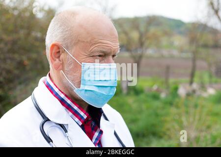 Portrait des älteren männlichen Arztes mit Uniform und chirurgischer Maske posiert im Freien Stockfoto