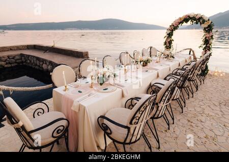 Nahaufnahme eines Festes am Esstisch für die Hochzeit. Ein Tisch steht am Strand mit Blick auf die Berge bei Sonnenuntergang. Metall geschmiedete Stühle, brennende Kerzen, Hochzeit Stockfoto