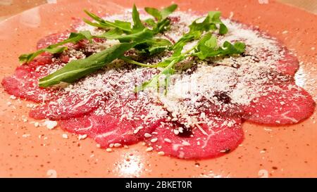 Nahaufnahme von Carpaccio Fleisch mit Parmesan und Rucola in einer Keramik großen orangefarbenen Platte auf einem Holztisch. Mit Olivenöl. Stockfoto