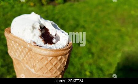 Eis in einer Waffelschale mit Schokoladenfüllung beißen. Im Sommer weißes Eis mit Karamell auf grünem Gras beißen. Stockfoto