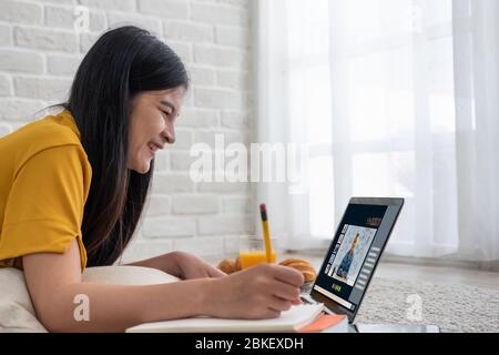 Asiatische weibliche Schüler Videokonferenz mit Lehrer und Freunde mit E-Learning-App auf Laptop-Computer zu Hause.Frau auf Teppich liegend.New normal le Stockfoto