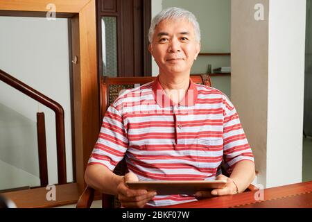 Porträt des glücklichen älteren chinesischen Mannes im gestreiften Hemd, der auf Tablet-Computer an seinem Schreibtisch zu Hause arbeitet Stockfoto
