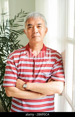 Ernst reifen chinesischen Mann zu Hause bleiben während der Lockdown, steht er am Fenster und Blick auf Kamera Stockfoto