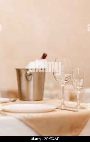 Vertikaler Rahmen der Tischeinstellung mit Gläsern, Teller, Messer, im Hintergrund Eimer Eis und Flasche Champagner. Stockfoto