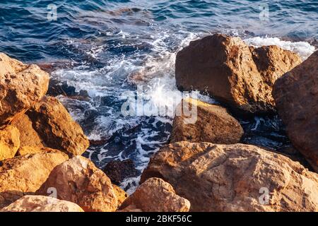 Blaue schäumende Wellen schlagen gegen große warme Steine, die an der Küste von der Sonne erleuchtet werden. Stockfoto